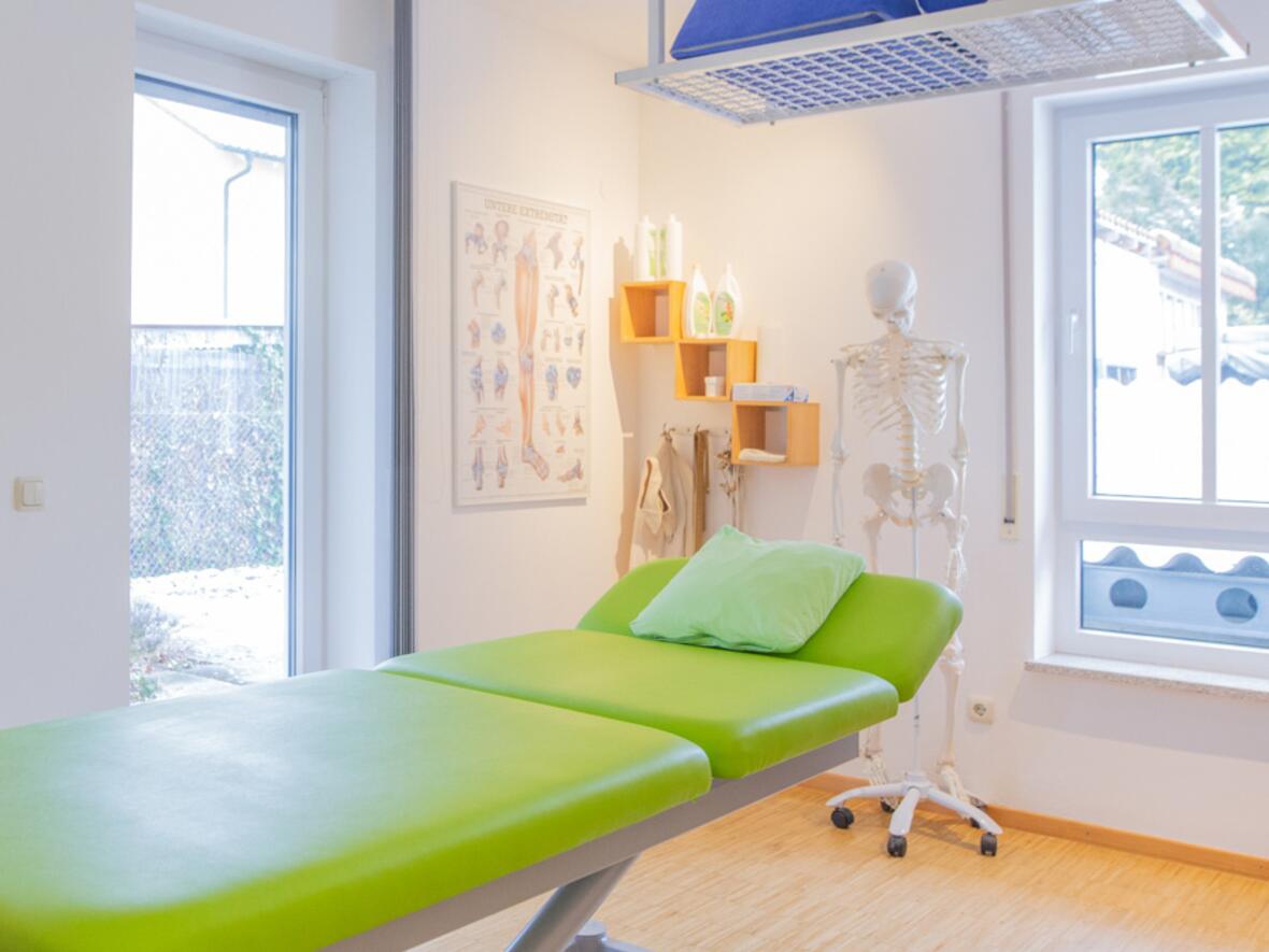 Behandlungsraum mit einem grünen Therapietisch, einem Skelett und medizinischen Poster an der Wand.