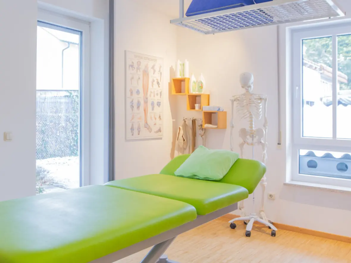 Behandlungsraum mit einem grünen Therapietisch, einem Skelett und medizinischen Poster an der Wand.