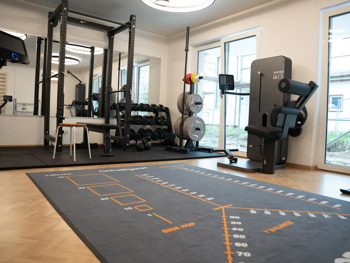 Fitnessraum mit Trainingsgeräten, Hanteln und Übungsteppich, große Fenster zur Außenansicht.