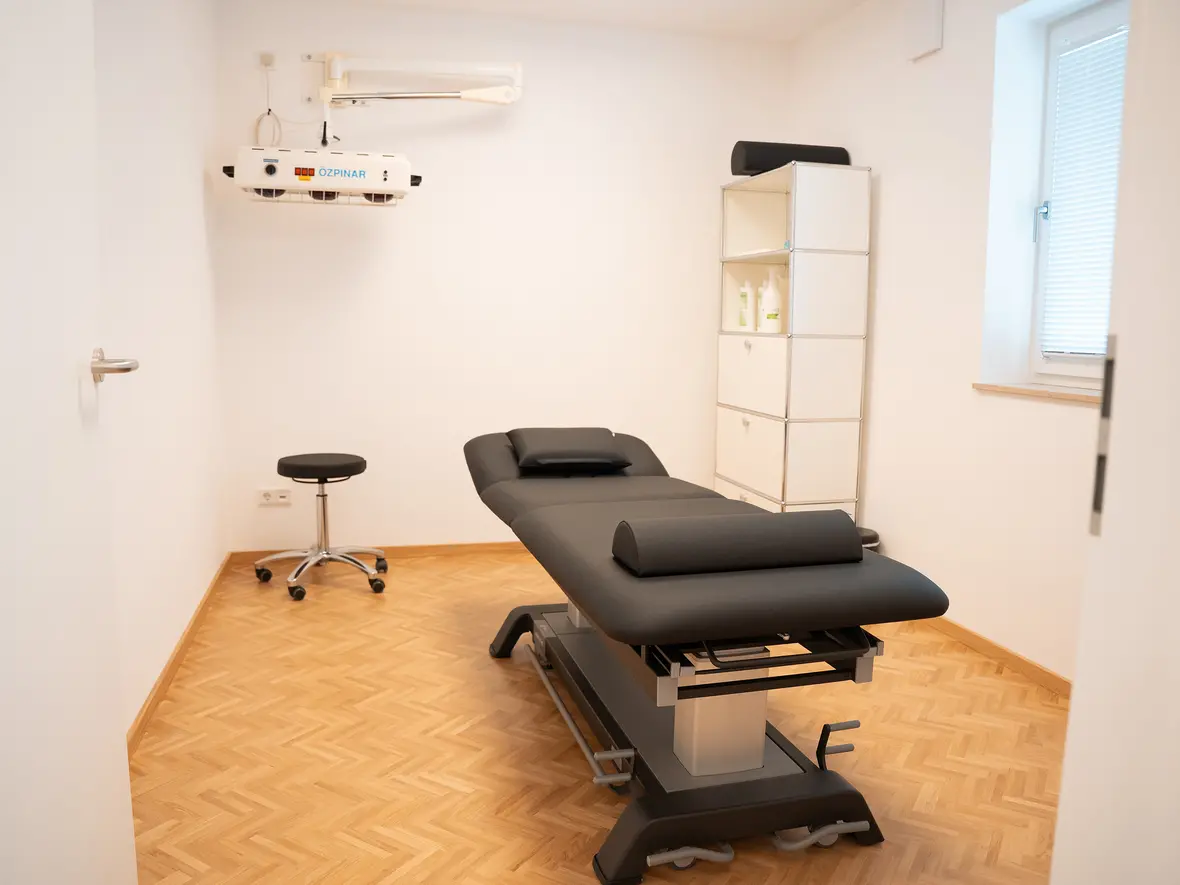 Therapieraum mit Behandlungsliege, Stuhl und Regal für medizinische Utensilien.