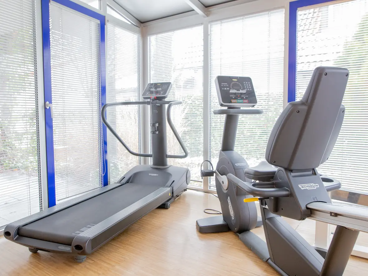 Moderner Trainingsraum der Physiotherapie Bartochek mit Laufband und Sitzergometer vor großen Glasfronten mit Jalousien.