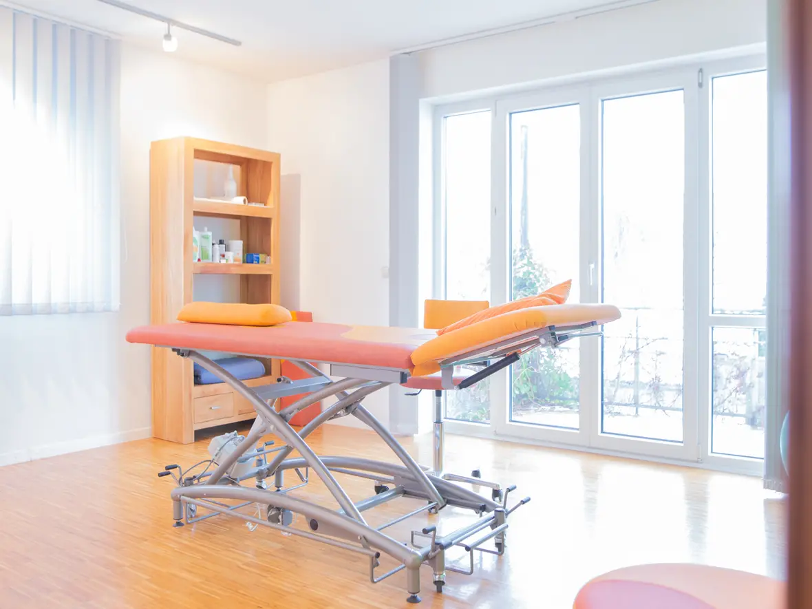 Freundlich eingerichteter Behandlungsraum der Physiotherapie Bartochek mit höhenverstellbarer orangefarbener Therapieliege und großem Fenster für natürliches Licht.