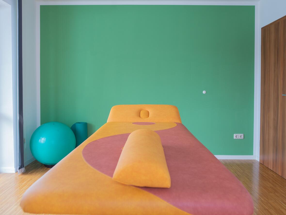 Moderner Behandlungsraum der Physiotherapie Bartochek mit grüner Wand, orangefarbener Therapieliege und Gymnastikball am Fenster.