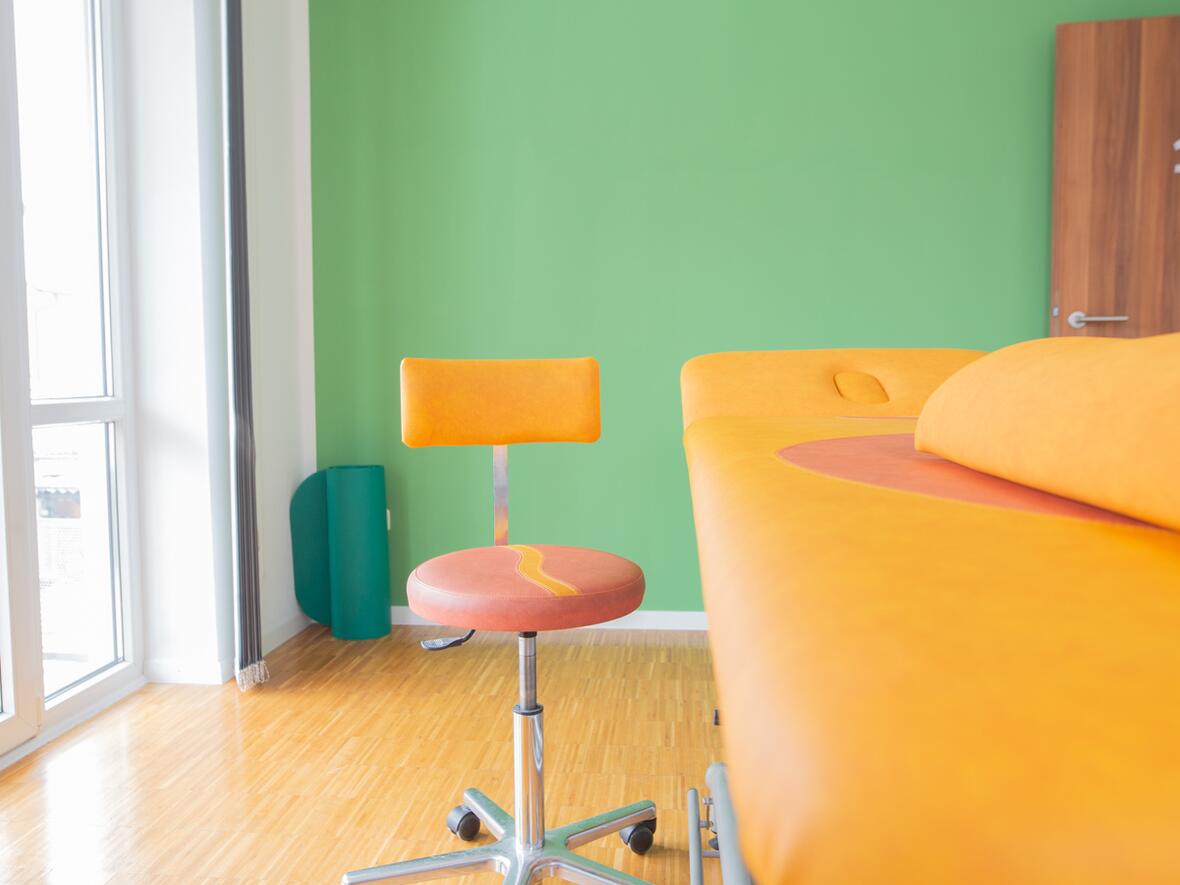 Freundlicher Behandlungsraum der Physiotherapie Bartochek mit orangefarbener Therapieliege, passendem Drehstuhl und grüner Wand, durch große Fenster gut belichtet.