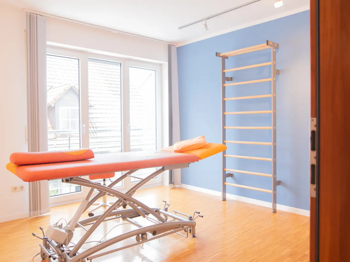 Heller Behandlungsraum der Physiotherapie Bartochek mit orangefarbener Therapieliege, blauer Wand und moderner Sprossenwand für Trainingsübungen.