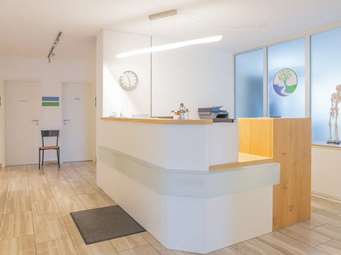 Heller Empfangsbereich der Physiotherapie Bartochek mit moderner Theke, Holz- und Glaselementen, Wanduhr und Praxislogo im Hintergrund.