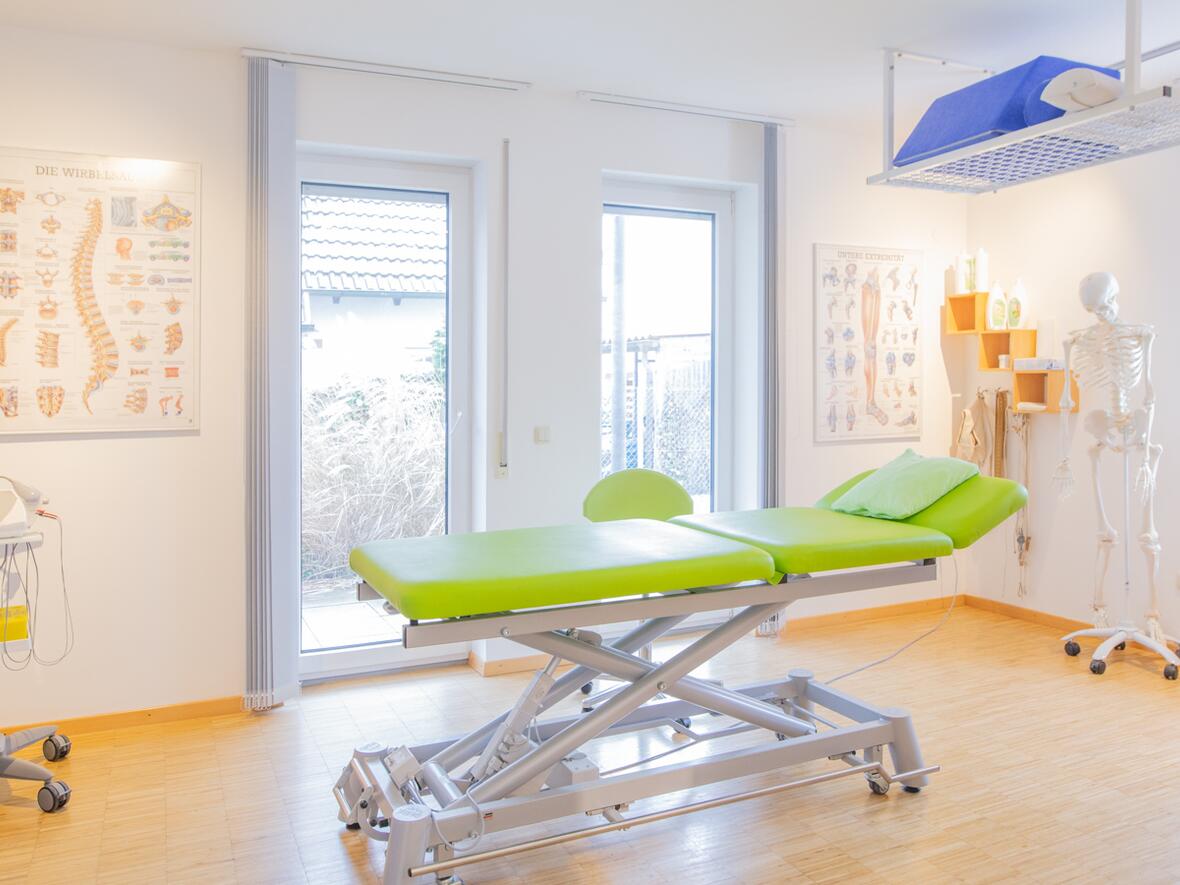 Heller Behandlungsraum der Physiotherapie Bartochek mit grüner Therapieliege, anatomischen Wandpostern und Therapiegeräten, freundlich und modern eingerichtet.