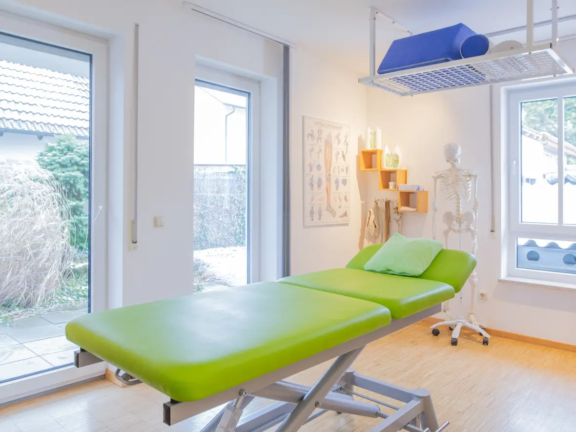 Lichtdurchfluteter Behandlungsraum der Physiotherapie Bartochek mit grüner Therapieliege, anatomischen Wandpostern und Skelettmodell vor großen Fenstern.