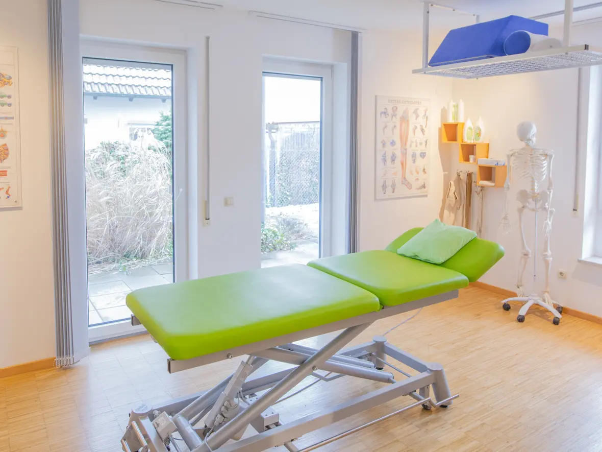 Heller Behandlungsraum der Physiotherapie Bartochek mit grüner Therapieliege, anatomischen Postern, Skelettmodell und großen Fenstern mit Blick nach draußen.