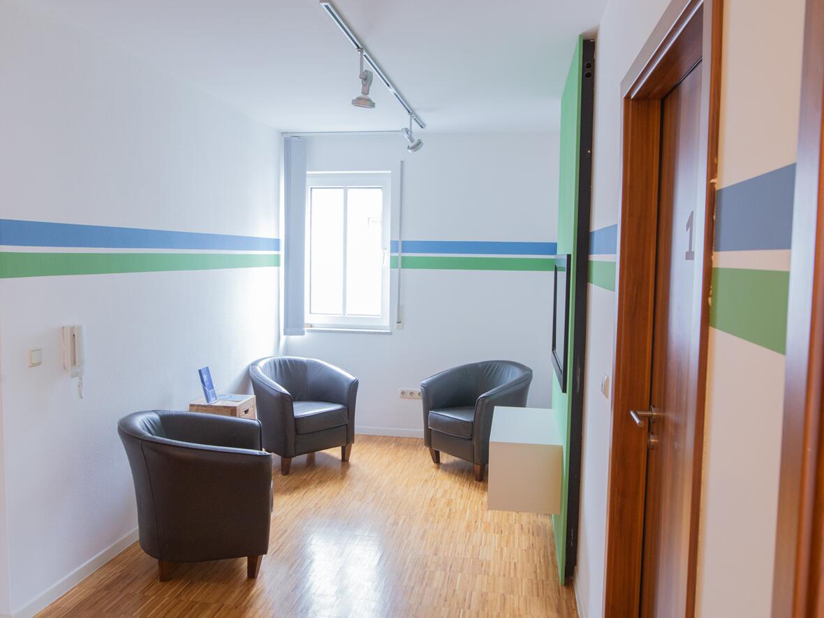 Heller Wartebereich der Physiotherapie Bartochek mit drei dunklen Ledersesseln, farbigen Wandstreifen in Grün und Blau sowie Holzboden.